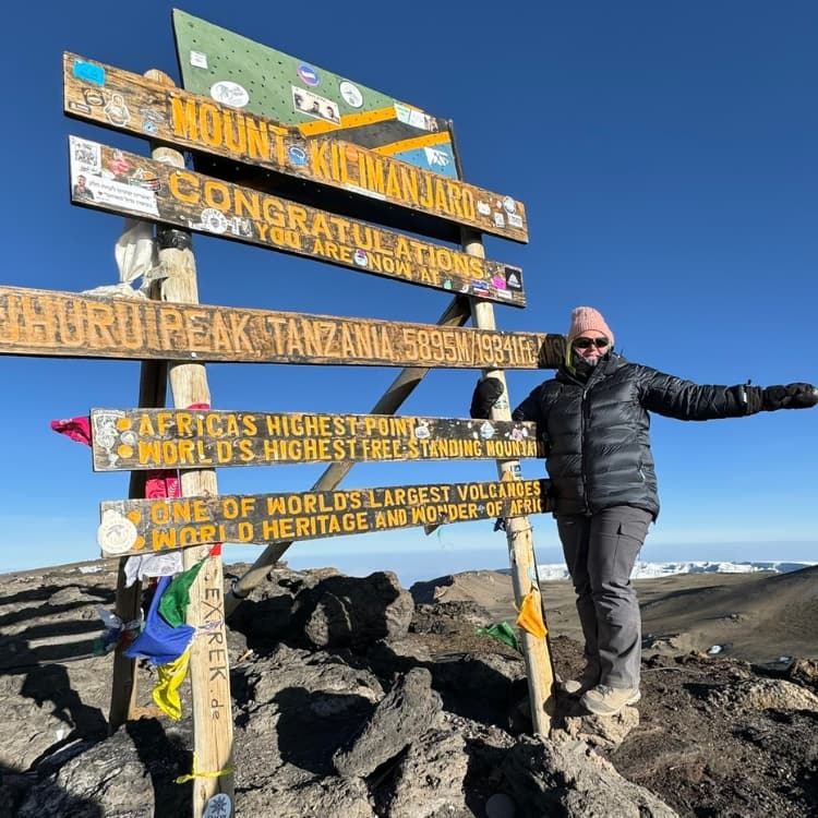 Climber at Uhuru Peak summit sign 5895 metres with retreating glacier visible behind on Kilimanjaro.
